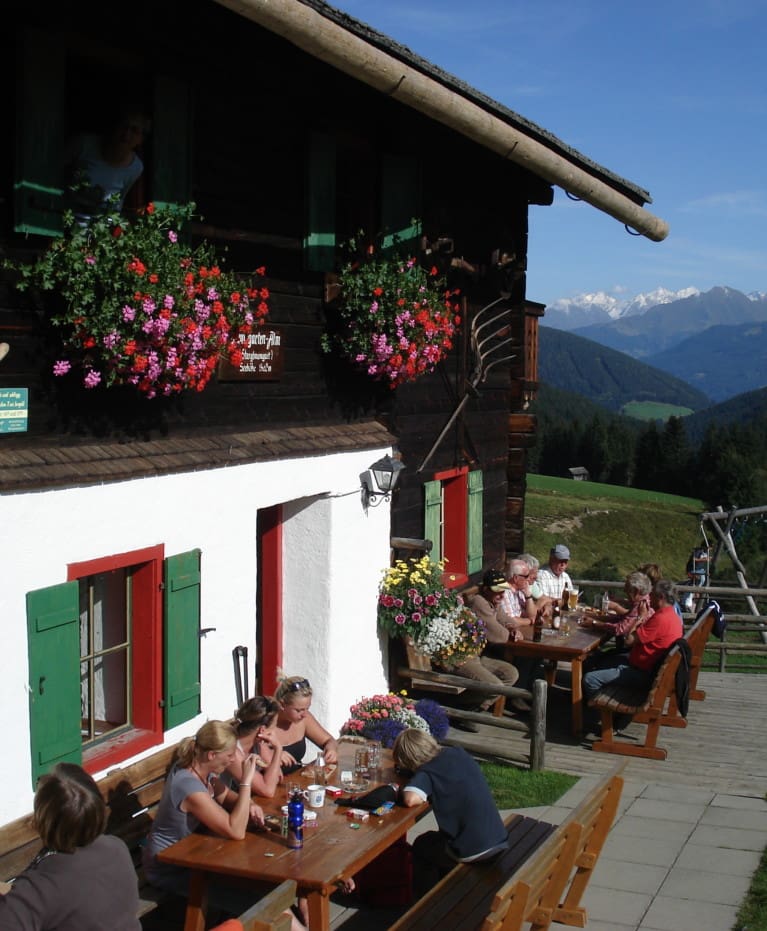 Die Menschen sitzen an hölzernen Picknicktischen vor einem rustikalen, mit bunten Blumen geschmückten Wanderhotel und genießen Speisen und Getränke in einer sonnigen Berglandschaft mit Blick auf schneebedeckte Gipfel und grüne Hügel.