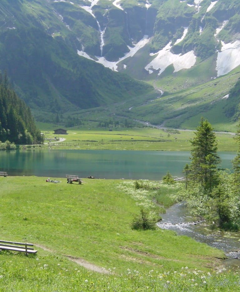 Ein friedlicher See, umgeben von grünem Gras, Bäumen und schneebedeckten Bergen. Im Vordergrund stehen Bänke an einem Bach, in der Ferne wandern Menschen unter einem klaren, sonnigen Himmel - perfekt für Gäste eines nahe gelegenen Wanderhotels.