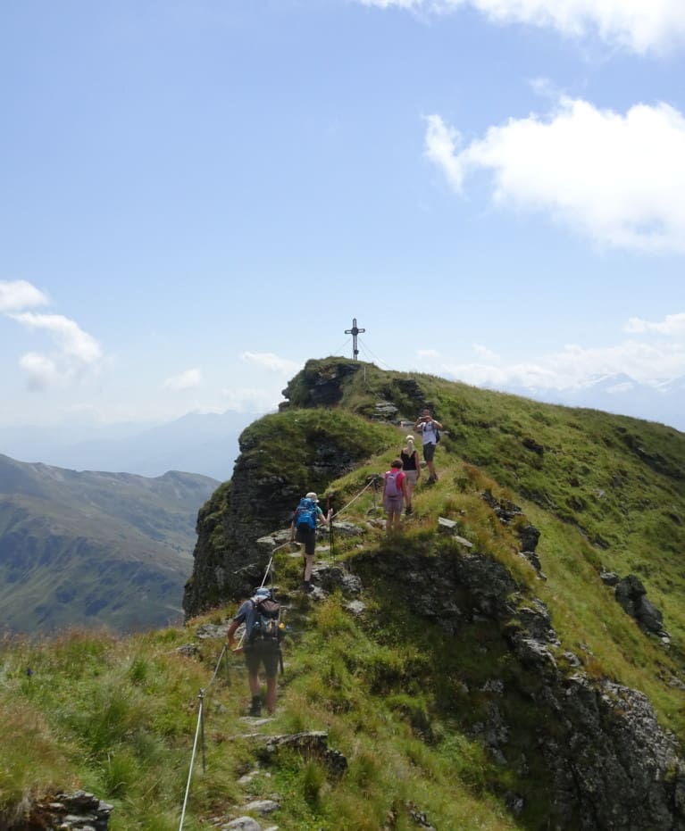 Eine Gruppe von Wanderern aus einem nahe gelegenen Wanderhotel wandert entlang eines schmalen, grasbewachsenen Bergrückens auf einen mit einem großen Kreuz markierten Gipfel zu, mit blauem Himmel und fernen Bergen im Hintergrund.