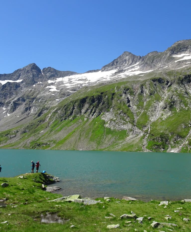 Eine Gruppe von Menschen steht am Rande eines türkisfarbenen Bergsees in der Nähe eines Wanderhotels, umgeben von felsigen, grünen Hängen und schneebedeckten Gipfeln unter einem klaren blauen Himmel.