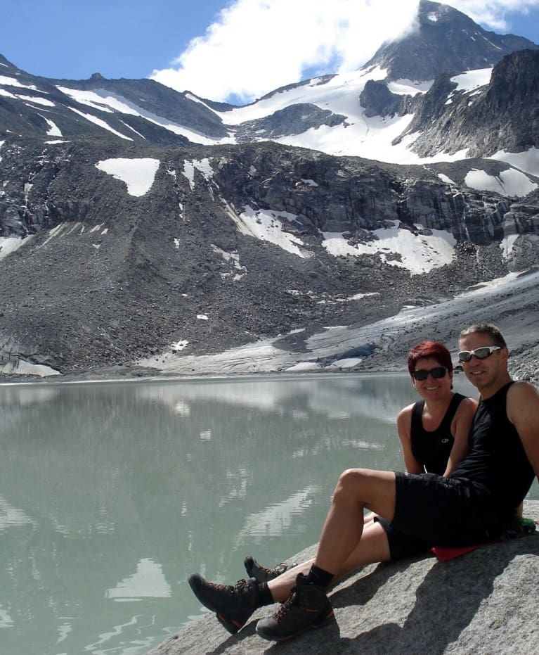 Zwei Personen in Wanderausrüstung sitzen auf einem Felsen an einem spiegelnden Bergsee und lächeln in die Kamera, während im Hintergrund schneebedeckte Gipfel und felsiges Gelände zu sehen sind - eine perfekte Wanderhotel-Atmosphäre. Eine dritte Person sitzt in der Nähe und schaut weg.