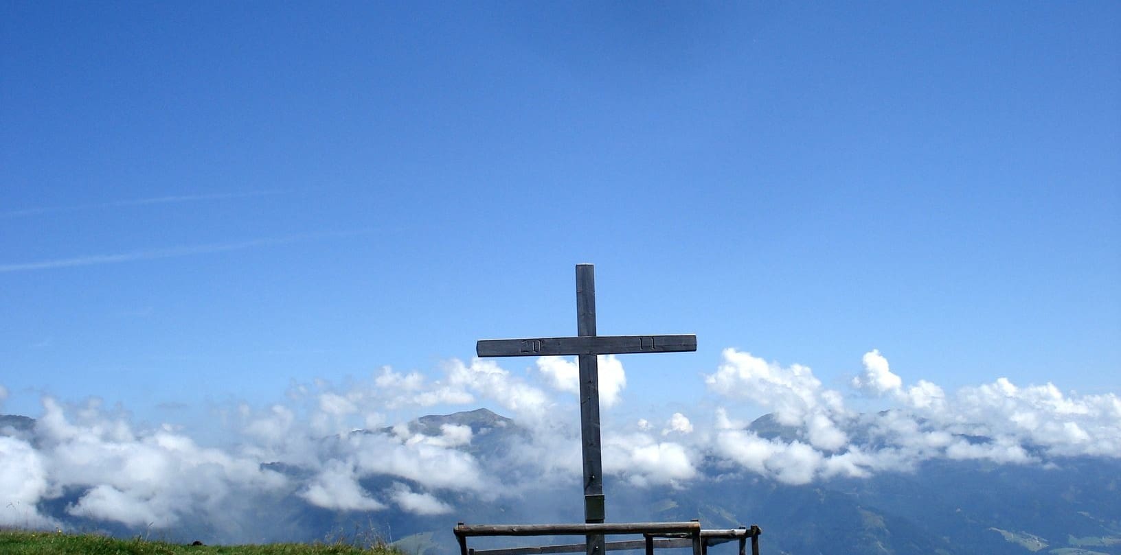Ein einfaches Holzkreuz steht auf der Poelsenalm Platte gegen einen strahlend blauen Himmel, umrahmt von schwebenden weißen Wolken und dem fernen Blick auf die nebelverhangenen Berge