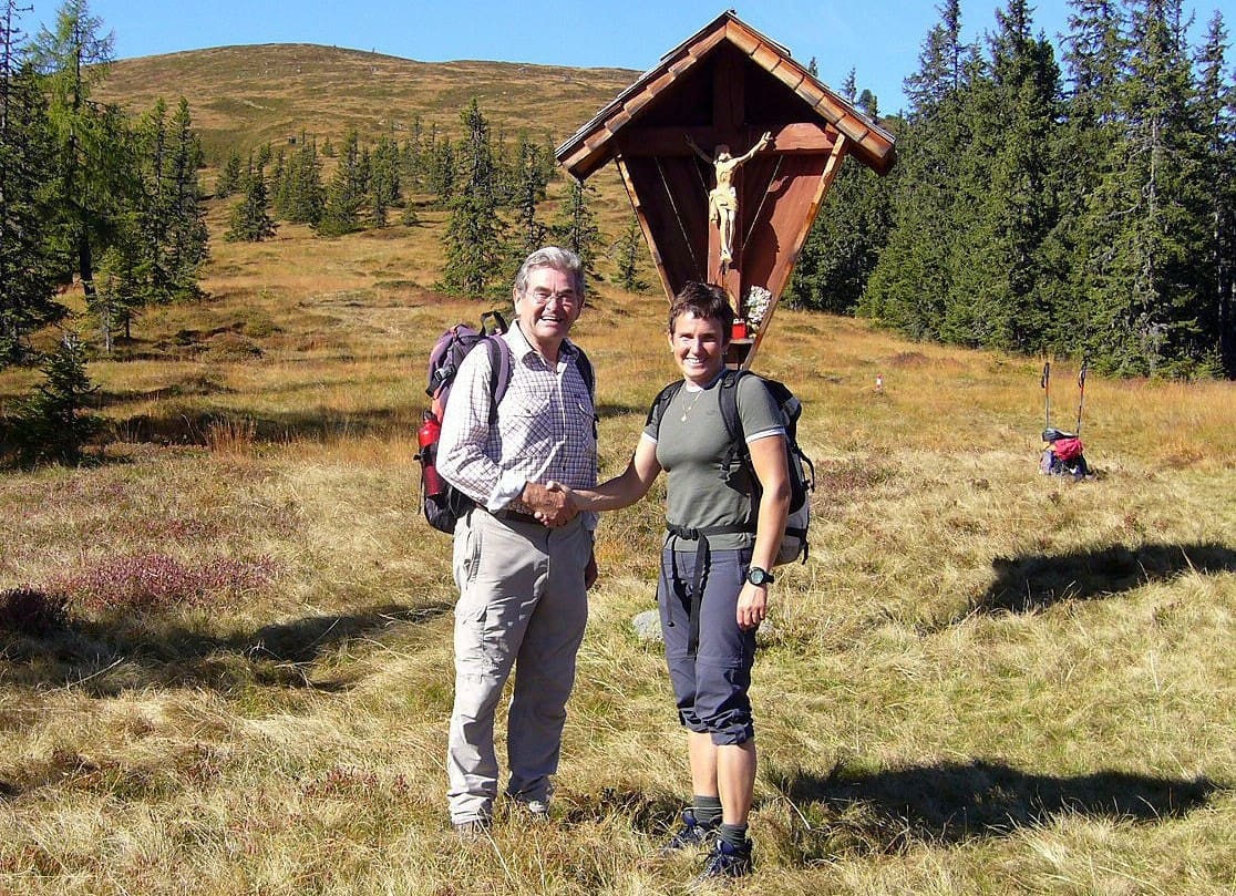 Zwei glückliche Wanderer geben sich vor einem traditionellen Wetterkreuz die Hand, das auf einer lichten Waldlichtung im Filzgebiet steht